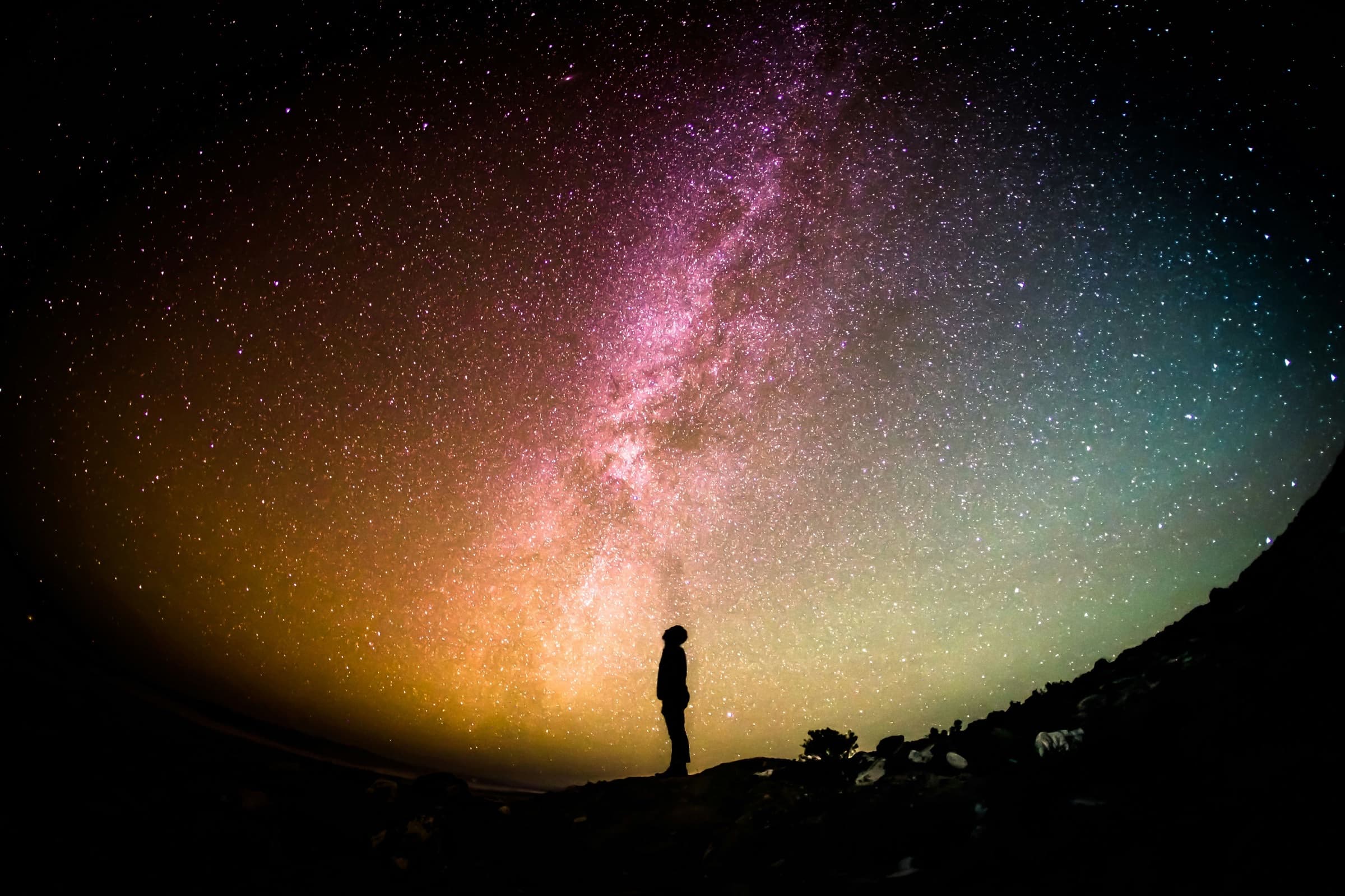 A lone figure standing on a ridge, looking up at the Milky Way arching across a star-filled sky
