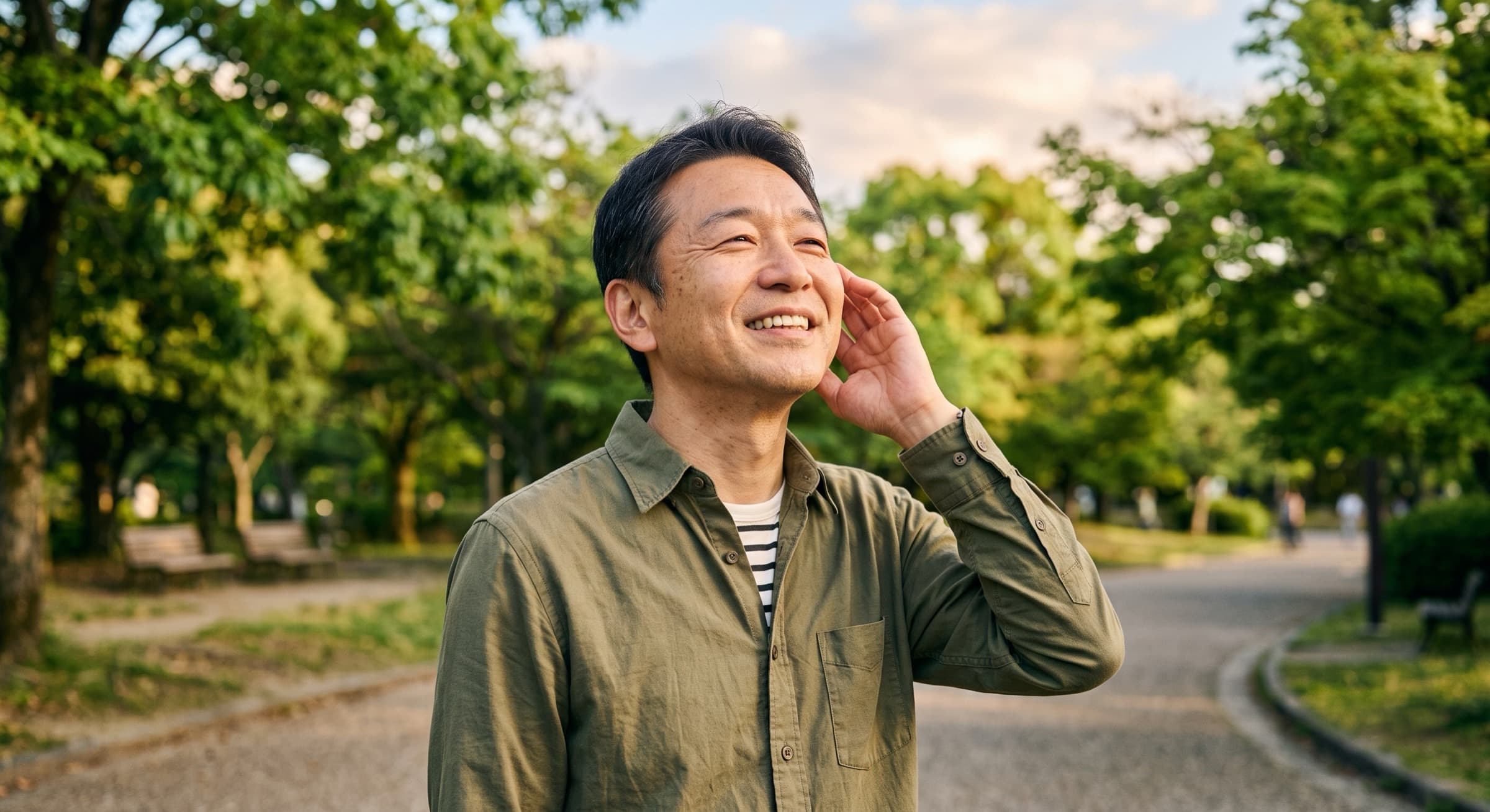 A man in a park cupping a hand behind his ear to listen, smiling and looking upward