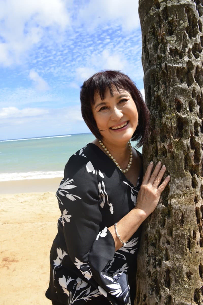 Michele Okimura on a beach in Hawaiʻi, leaning against a palm trunk with the ocean behind her