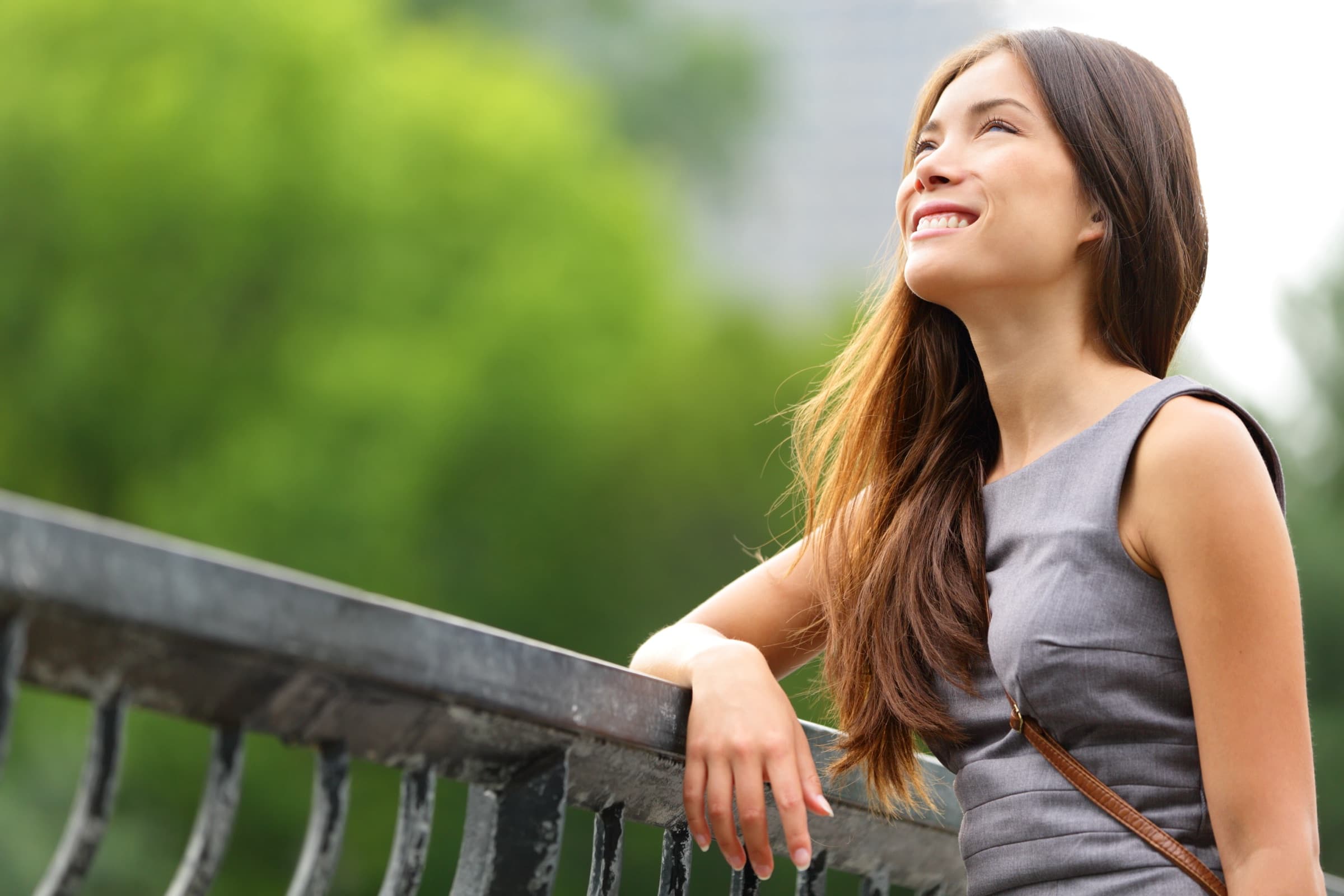 A woman leaning on a park railing, smiling as she looks up, sunlit green trees blurred behind her